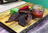 Dark reddish-brown beef jerky strips arranged on a wooden cutting board, served with a small glass jar of red chili sauce, a red chili pepper, and mint leaves.